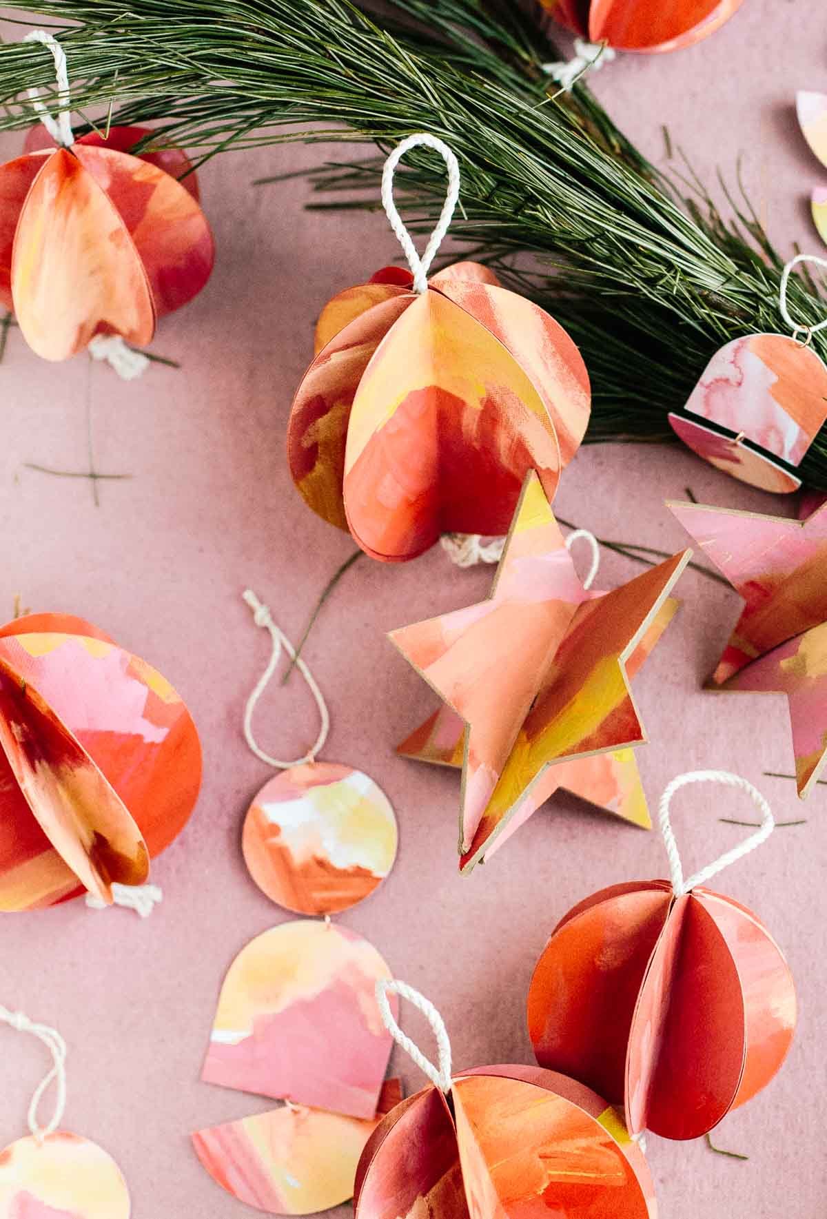 Christmas ornaments made of paper in various shapes laid out on a pink background with pine tree trimmings