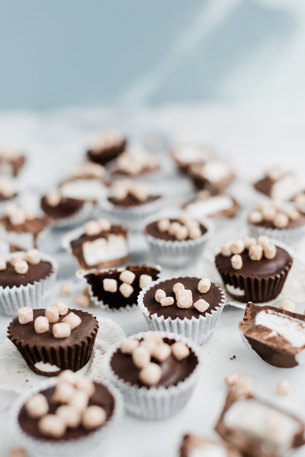 hot cocoa candy cups with chocolate and tiny marshmallows on top, scattered on a marble countertop with light blue background