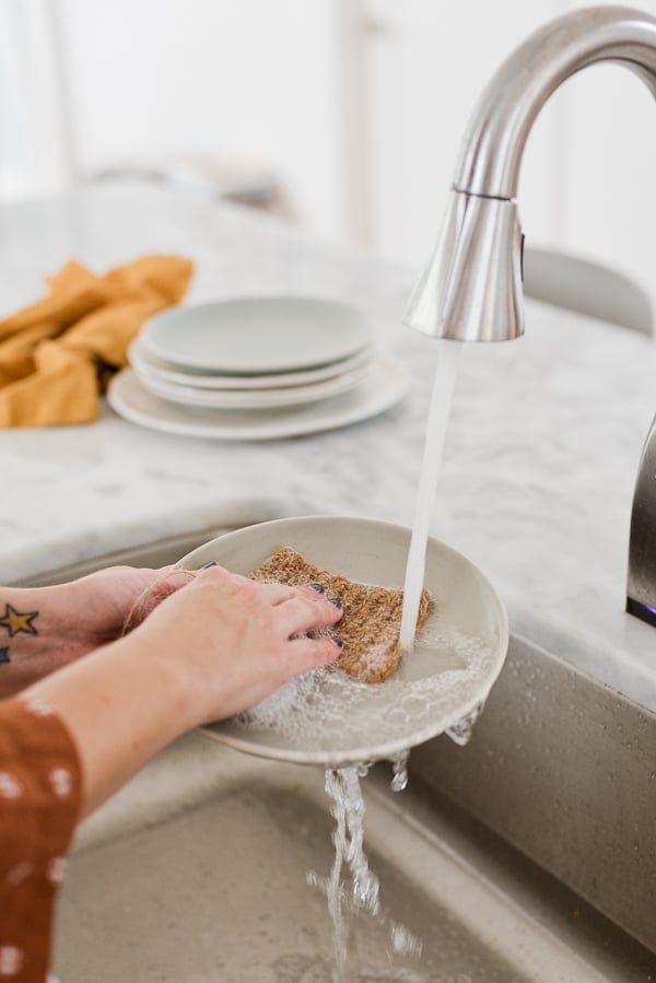 Running water over a dish in the sink, using a natural jute dish scrubber.