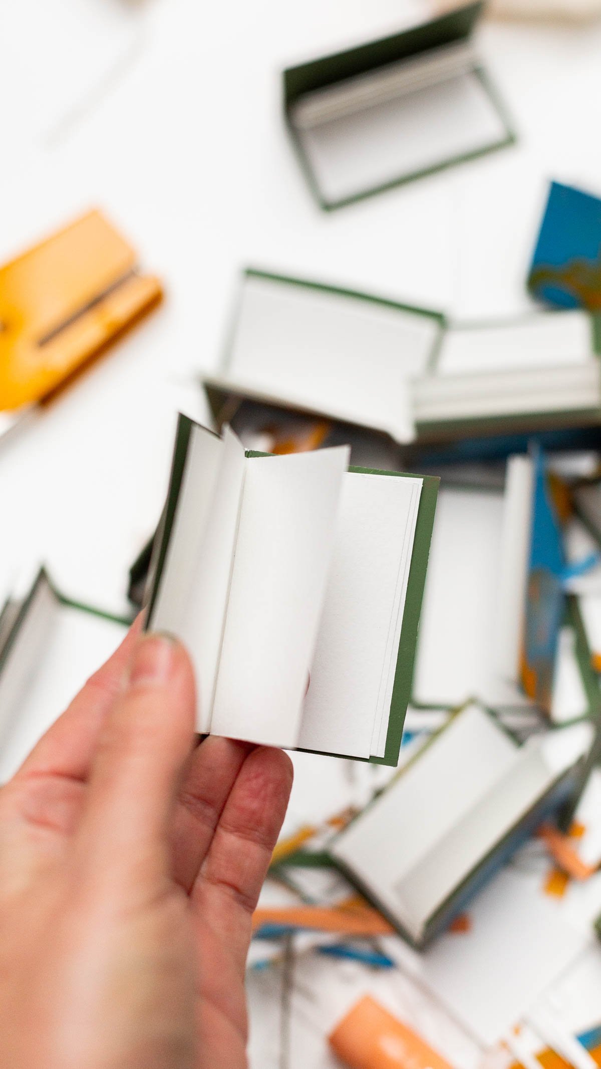 person holding tiny homemade notebook in hand with mess of supplies behind 