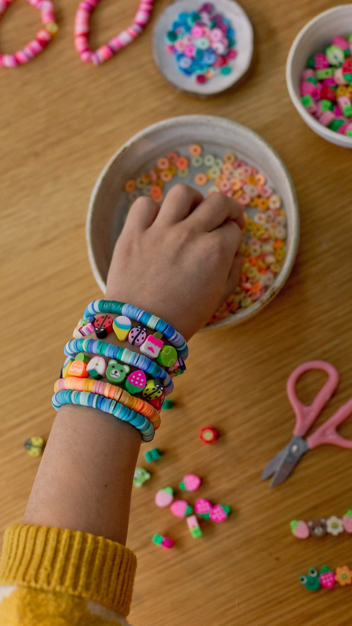child's hand reaching into a bowl of clay beads, who is wearing a lot of clay bead bracelets on his wrist