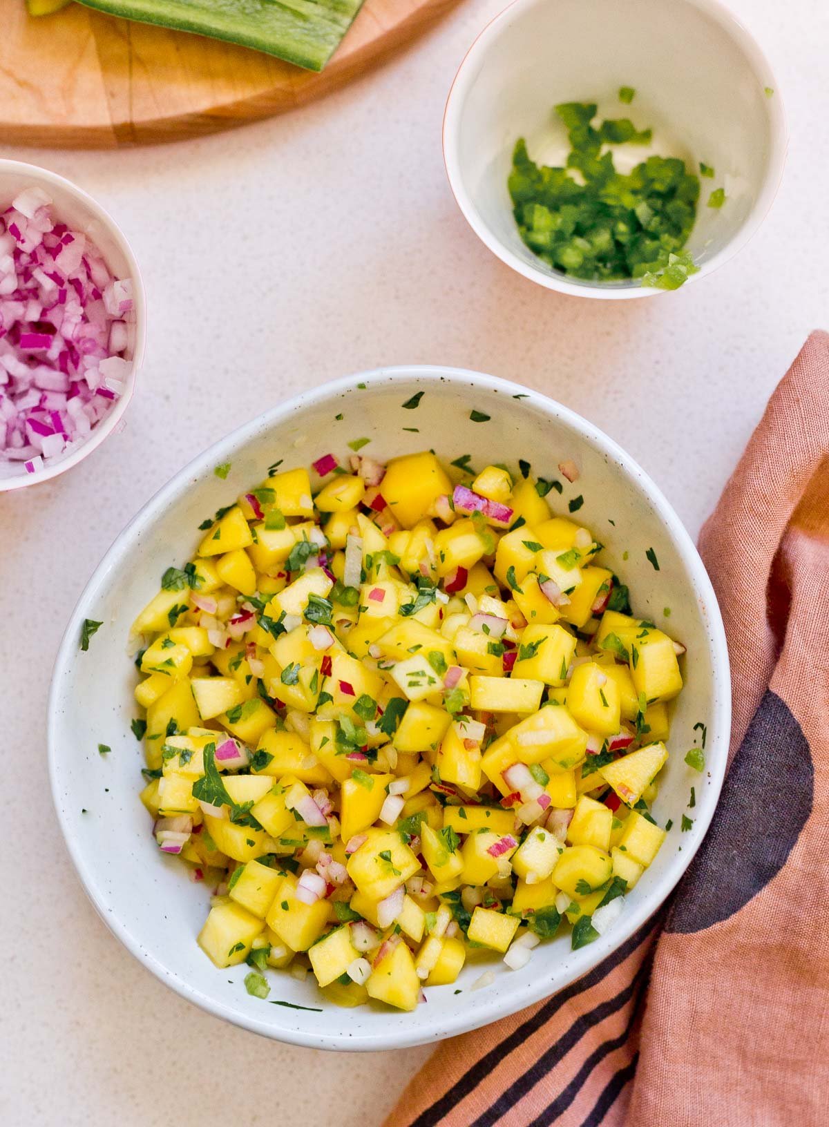 mango salsa in white bowl on table, surrounded by ingredients 