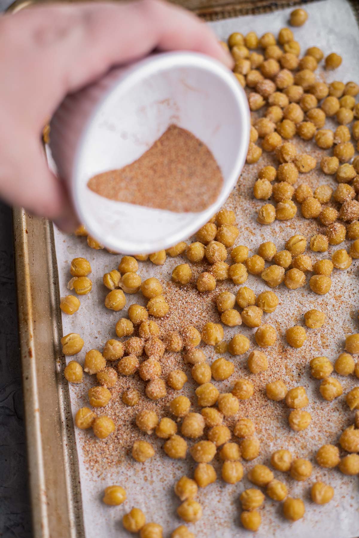 pouring spices onto roasted chickpeas that are on a baking sheet