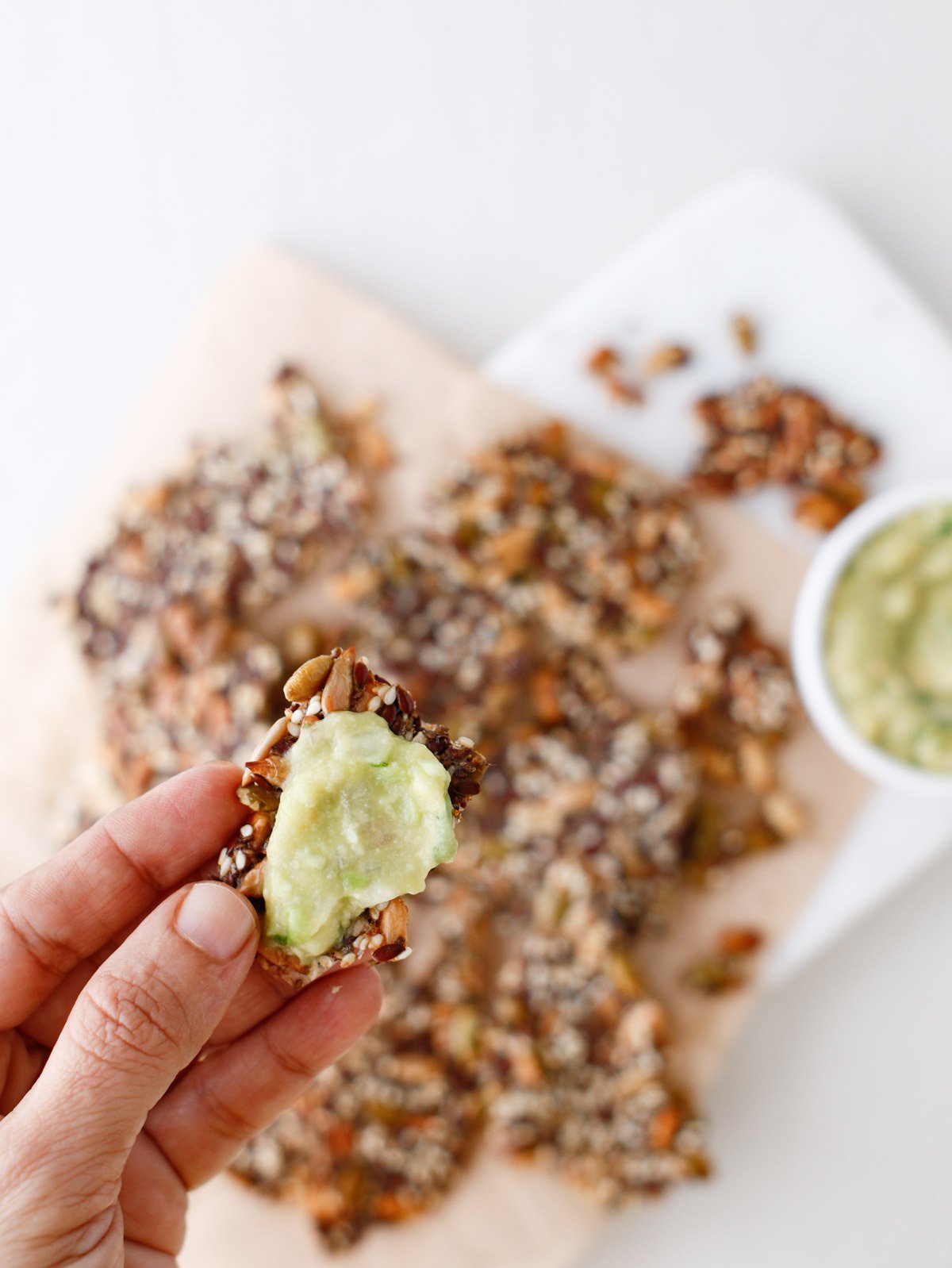 woman's hand holding small seed cracker with green veggie dip on it