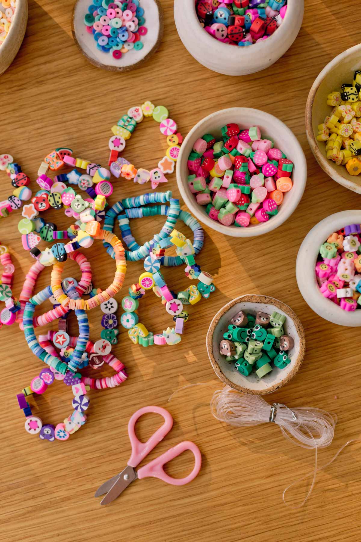 bowls of beads and clay bead bracelets laying out on wood surface with scissors and other supplies