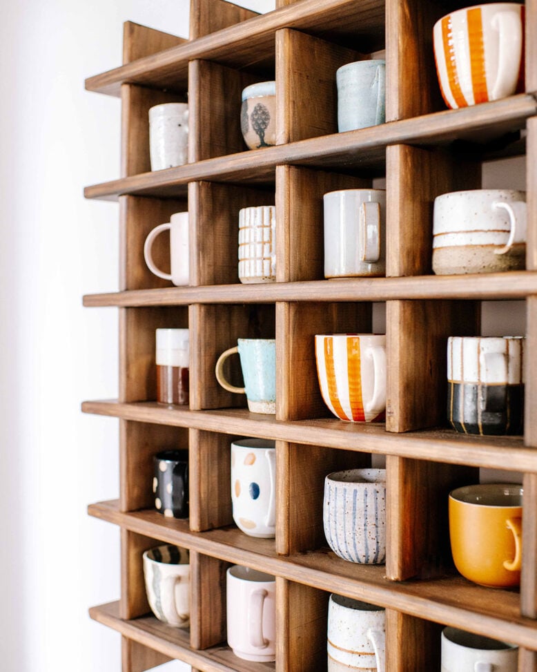 wood wall shelf with many small boxes, hanging on the wall, with handmade ceramic mugs inside each square
