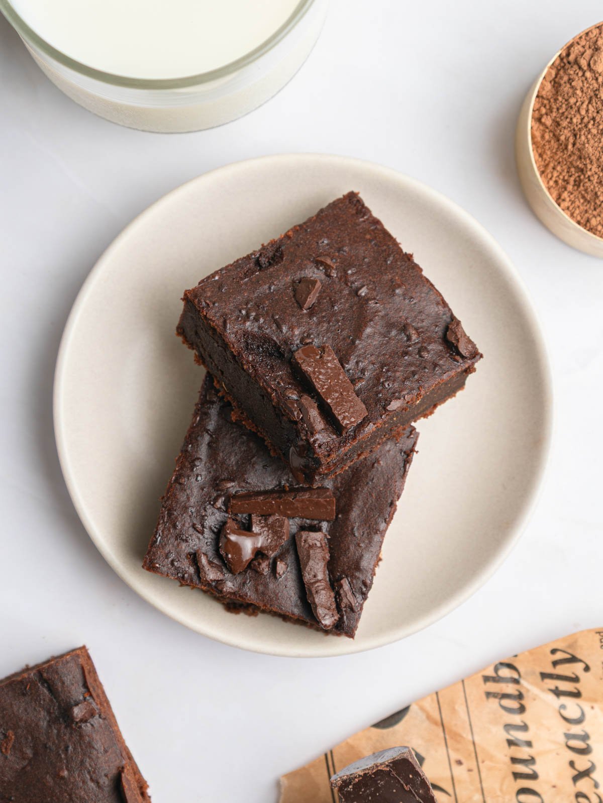 two square slices of brownie on a white plate with milk and cocoa powder