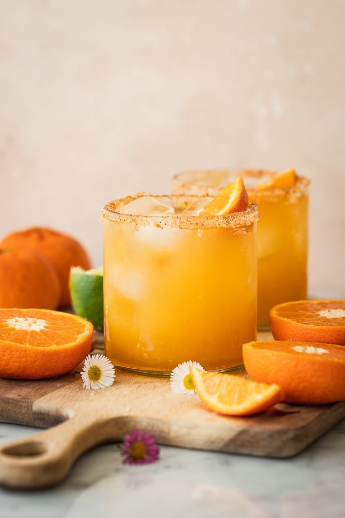 orange margaritas in short glasses with orange garnish, sitting on wood cutting board in a kitchen